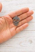 Silver-coloured metal skull connector with raised dotted detailing and attachment loops at the top and bottom, resting on a hand.