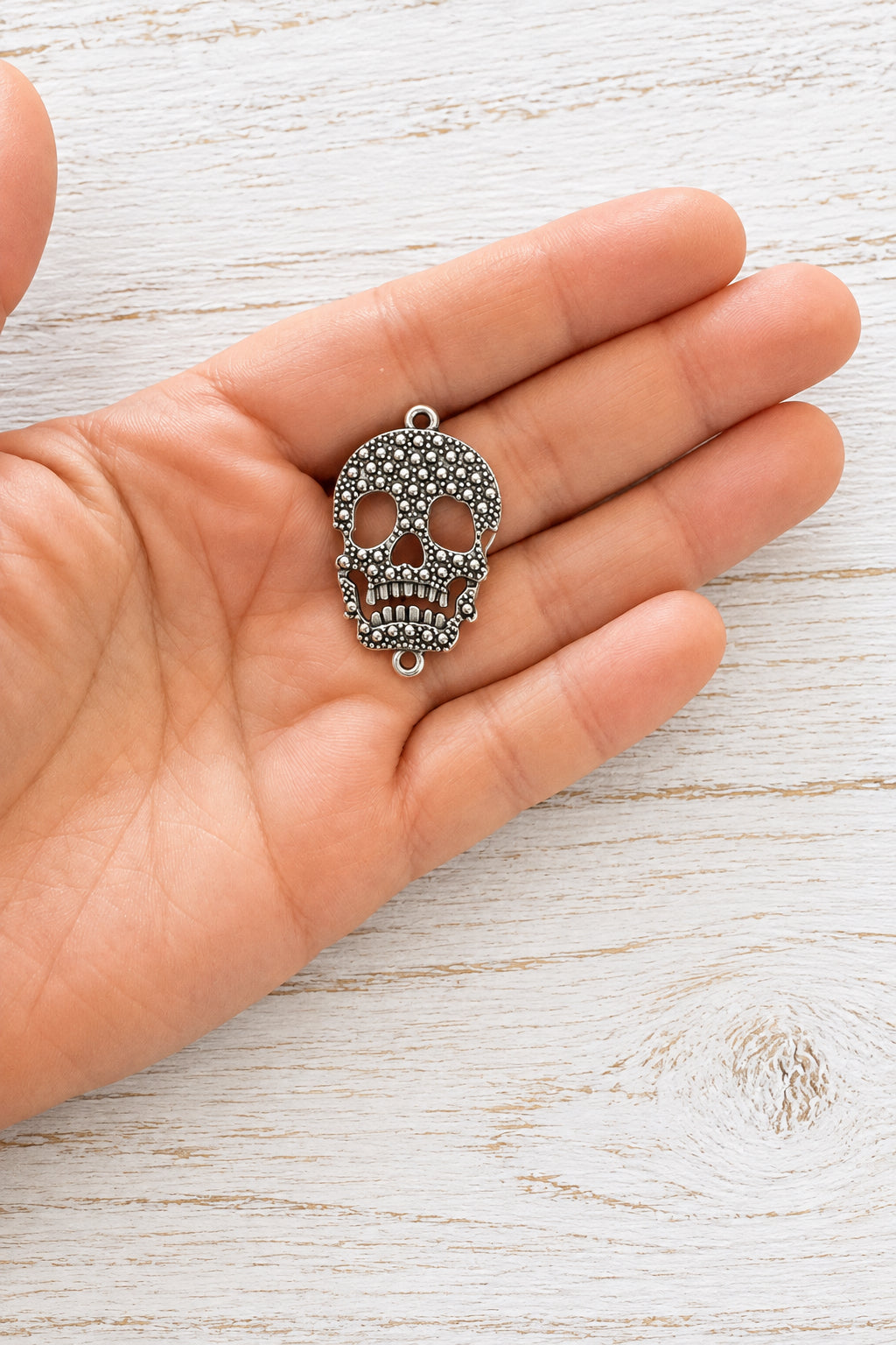 Silver-coloured metal skull connector with raised dotted detailing and attachment loops at the top and bottom, resting on a hand.