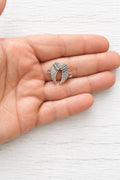 Silver-coloured metal angel wing connector with detailed feathers and two attachment loops, shown resting on a hand.