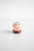 A small pink pig figurine standing on a brown base, topped with a pink macaron and green frosting, placed inside a pink tray.