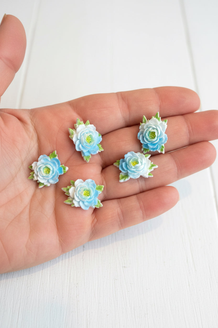 Small soft blue fabric flowers with green leaves held in a hand to show size, craft embellishments pack of 50
