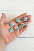 Small soft blue fabric flowers with green leaves held in a hand to show size, craft embellishments pack of 50