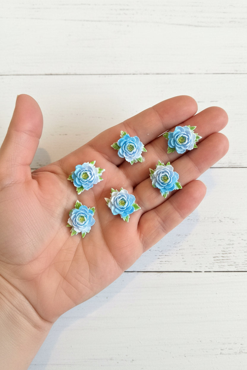 Small soft blue fabric flowers with green leaves held in a hand to show size, craft embellishments pack of 50